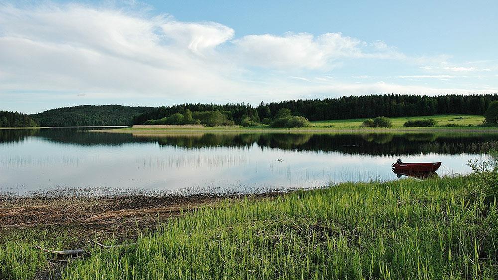 Innsjø med liten jolle i kanten, omringet av grønt gress - Klikk for stort bilde